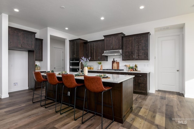 kitchen with a kitchen island with sink, visible vents, light countertops, under cabinet range hood, and dark wood-type flooring