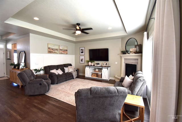 living room with recessed lighting, dark wood-style flooring, a ceiling fan, a lit fireplace, and a tray ceiling