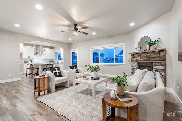 living area with light wood finished floors, baseboards, ceiling fan, a stone fireplace, and arched walkways