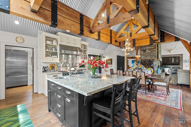 kitchen with a kitchen island with sink, wood finished floors, dark cabinetry, open shelves, and stainless steel built in refrigerator