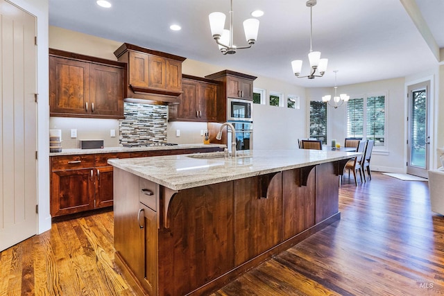 kitchen with a chandelier, a sink, appliances with stainless steel finishes, dark wood-type flooring, and an island with sink
