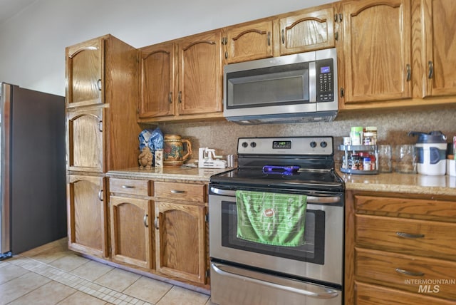 kitchen featuring light tile patterned floors, brown cabinetry, decorative backsplash, light stone counters, and appliances with stainless steel finishes