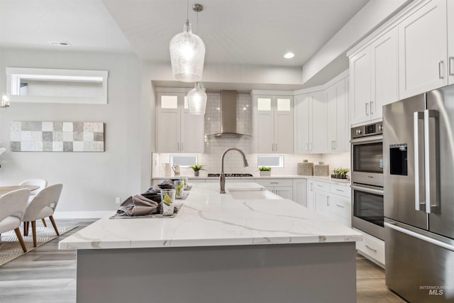 kitchen with stainless steel appliances, white cabinets, wall chimney range hood, and an island with sink