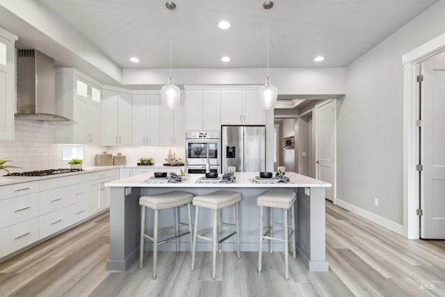 kitchen featuring appliances with stainless steel finishes, white cabinetry, wall chimney range hood, and an island with sink