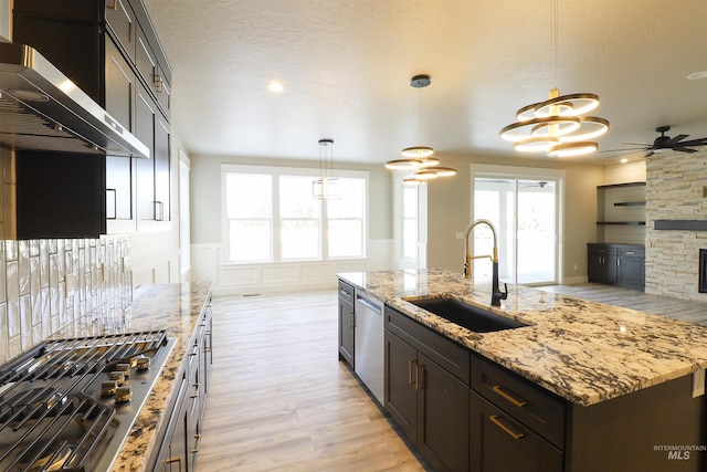 kitchen featuring sink, ventilation hood, hanging light fixtures, appliances with stainless steel finishes, and a kitchen island with sink