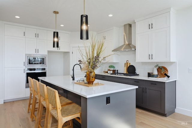 kitchen featuring wall chimney range hood, tasteful backsplash, white cabinetry, a kitchen island with sink, and stainless steel appliances