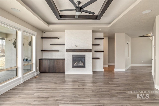 unfurnished living room with a glass covered fireplace, a tray ceiling, wood finished floors, and a ceiling fan