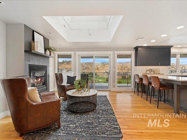 living area with a skylight, a glass covered fireplace, light wood-type flooring, a tray ceiling, and visible vents