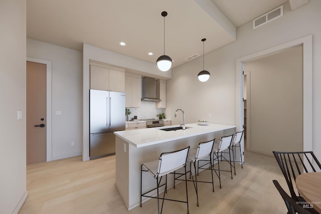 kitchen with built in refrigerator, decorative backsplash, wall chimney exhaust hood, sink, and decorative light fixtures