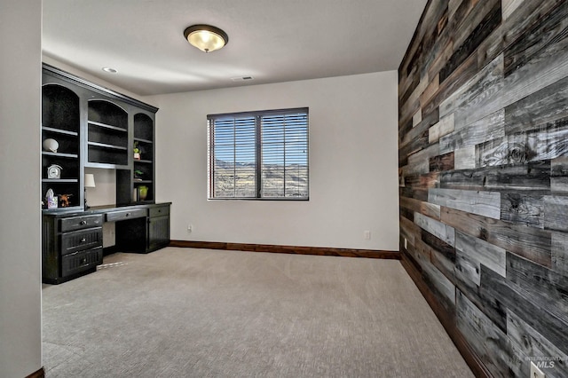 unfurnished office featuring wooden walls, visible vents, baseboards, built in desk, and light colored carpet