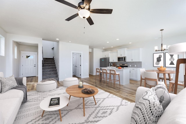 living area with recessed lighting, ceiling fan with notable chandelier, baseboards, stairs, and light wood-style floors