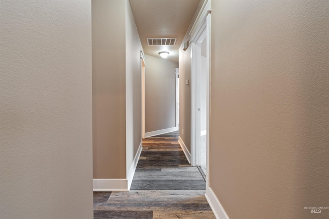 corridor with dark wood-type flooring, visible vents, a textured wall, and baseboards