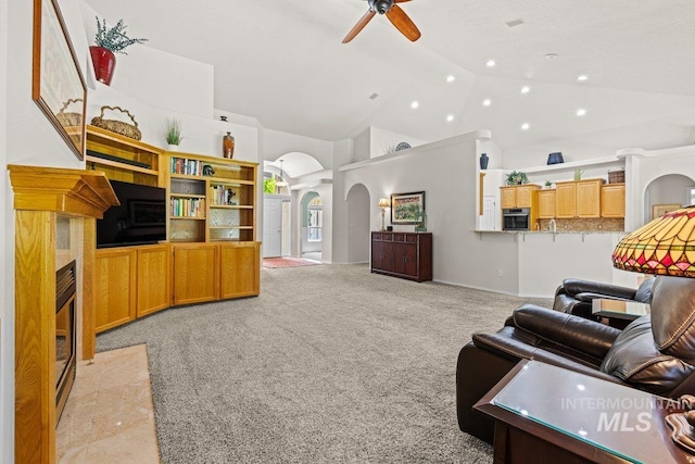 living room with arched walkways, a ceiling fan, a tile fireplace, high vaulted ceiling, and light colored carpet
