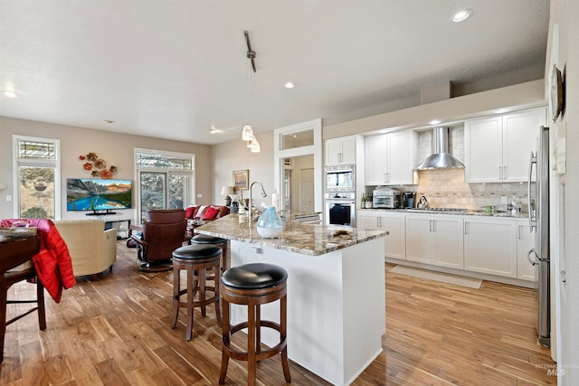 kitchen featuring decorative light fixtures, white cabinetry, a kitchen island with sink, stainless steel appliances, and wall chimney range hood