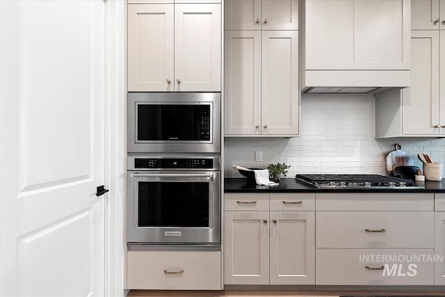 kitchen featuring white cabinets, dark countertops, decorative backsplash, custom range hood, and appliances with stainless steel finishes