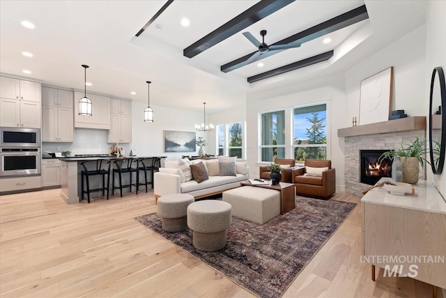 living room with ceiling fan with notable chandelier, a stone fireplace, light hardwood / wood-style flooring, and a raised ceiling