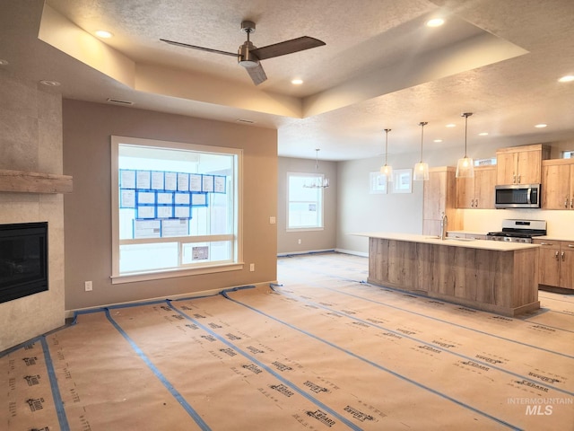 kitchen with a tile fireplace, pendant lighting, a raised ceiling, stainless steel appliances, and a center island with sink