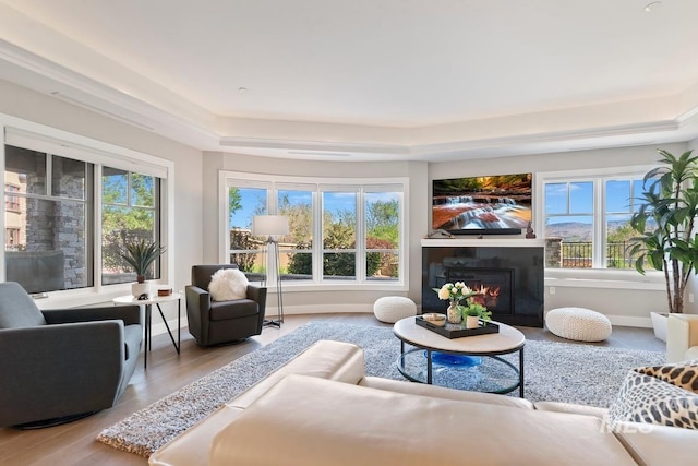 living room featuring baseboards, a tray ceiling, a lit fireplace, and wood finished floors