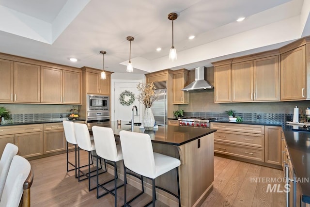 kitchen featuring a kitchen bar, a sink, dark countertops, built in appliances, and wall chimney range hood