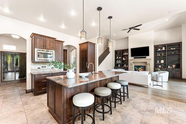 kitchen featuring arched walkways, stainless steel microwave, ceiling fan, dark stone countertops, and a glass covered fireplace
