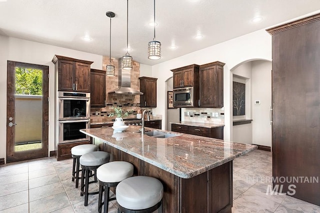 kitchen featuring appliances with stainless steel finishes, wall chimney exhaust hood, arched walkways, a sink, and decorative backsplash