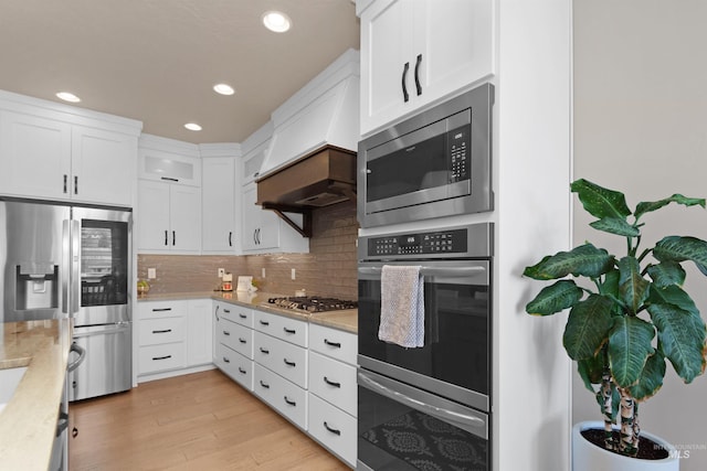 kitchen featuring custom exhaust hood, stainless steel appliances, decorative backsplash, white cabinets, and light wood-type flooring