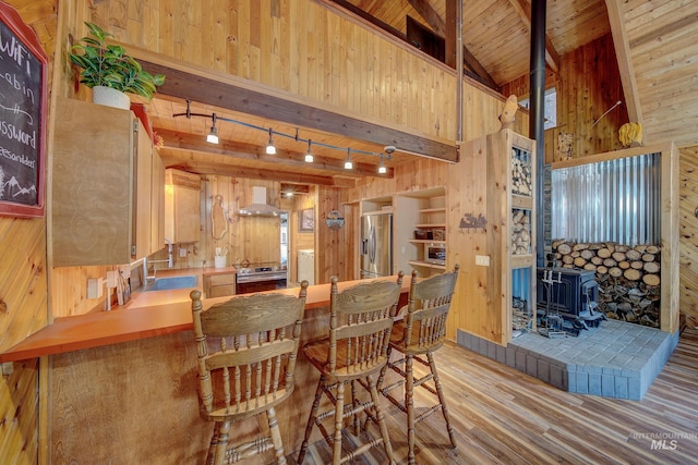 dining space with wood walls, a wood stove, rail lighting, beam ceiling, and light hardwood / wood-style flooring