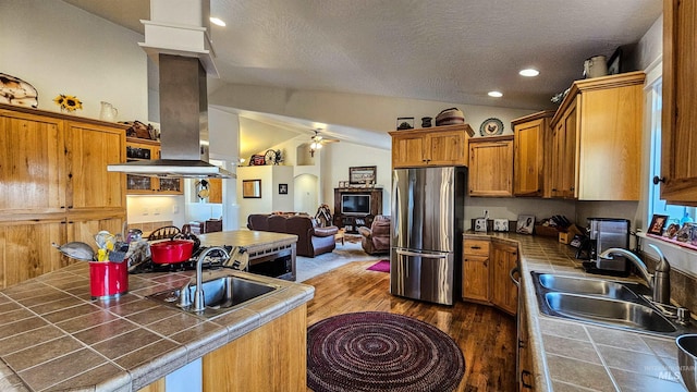 kitchen featuring vaulted ceiling, sink, stainless steel refrigerator, tile counters, and island range hood