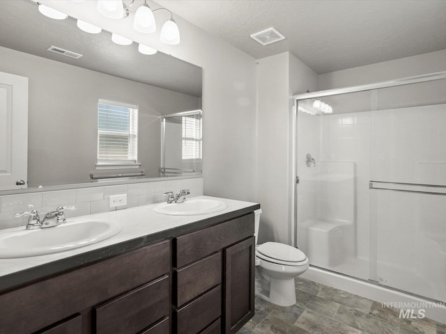 bathroom featuring a stall shower, toilet, double vanity, decorative backsplash, and stone finish floors