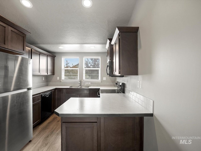 kitchen featuring appliances with stainless steel finishes, a peninsula, a sink, dark brown cabinets, and light wood-type flooring