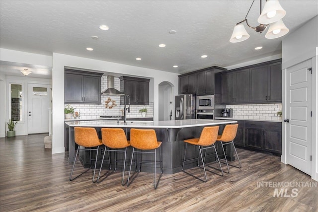 kitchen featuring dark wood-type flooring, wall chimney range hood, a spacious island, stainless steel appliances, and light countertops