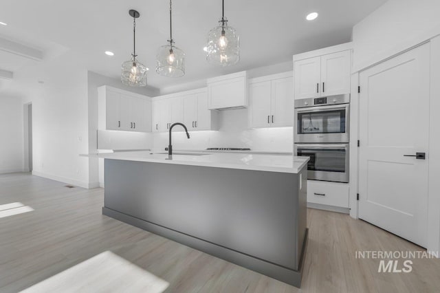 kitchen with stainless steel double oven, tasteful backsplash, light wood-type flooring, and a kitchen island with sink