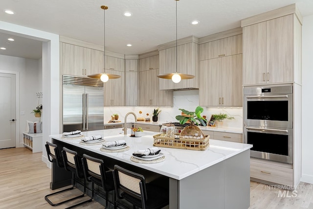 kitchen with a kitchen island with sink, a kitchen breakfast bar, stainless steel appliances, light stone countertops, and light wood-type flooring