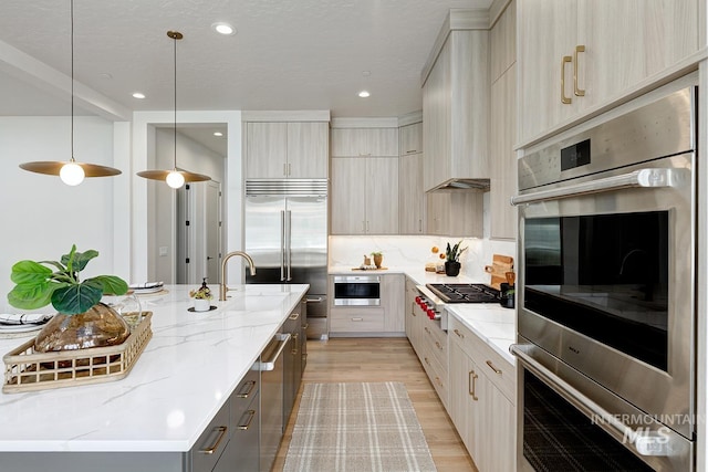 kitchen featuring appliances with stainless steel finishes, a kitchen island with sink, light stone countertops, light wood-type flooring, and pendant lighting