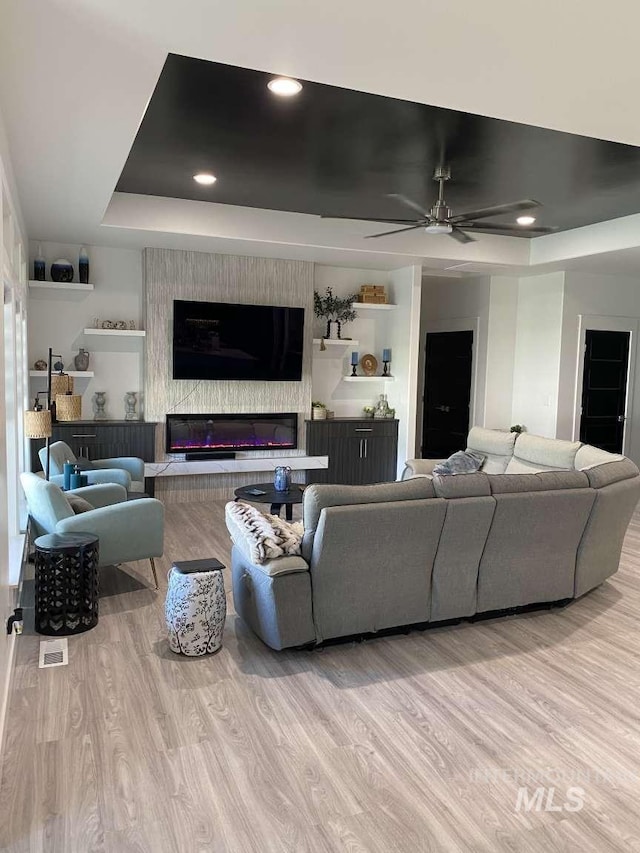 living room featuring a tray ceiling, ceiling fan, a fireplace, and light wood-type flooring