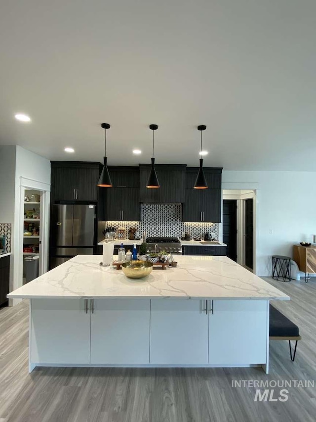 kitchen with tasteful backsplash, stainless steel fridge, decorative light fixtures, a kitchen island with sink, and light wood-type flooring