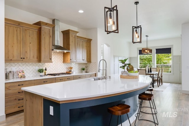 kitchen with a large island, tasteful backsplash, wall chimney exhaust hood, a sink, and light wood-style flooring