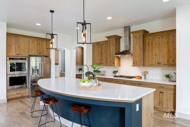 kitchen featuring appliances with stainless steel finishes, light wood-style flooring, wall chimney exhaust hood, backsplash, and a center island with sink