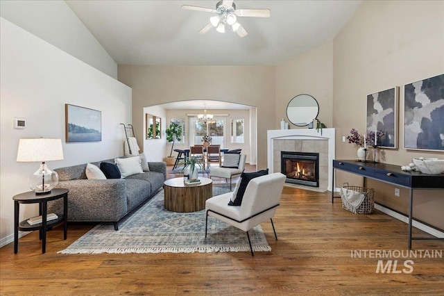 living room featuring arched walkways, wood finished floors, high vaulted ceiling, a tile fireplace, and ceiling fan with notable chandelier