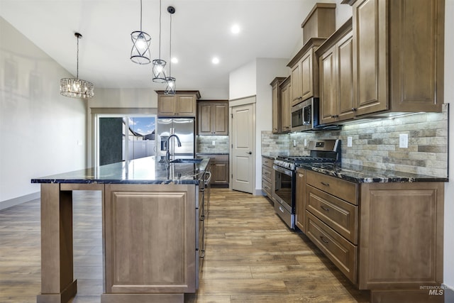 kitchen with decorative light fixtures, wood-type flooring, sink, stainless steel appliances, and a center island with sink