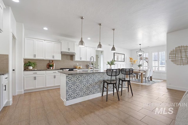 kitchen with a sink, backsplash, white cabinetry, a center island with sink, and dark stone countertops
