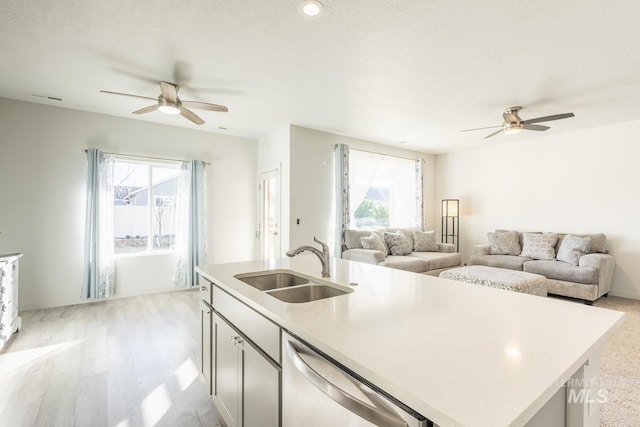 kitchen with sink, a kitchen island with sink, stainless steel dishwasher, ceiling fan, and light wood-type flooring