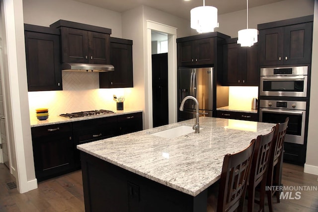 kitchen featuring a sink, light stone counters, under cabinet range hood, backsplash, and appliances with stainless steel finishes