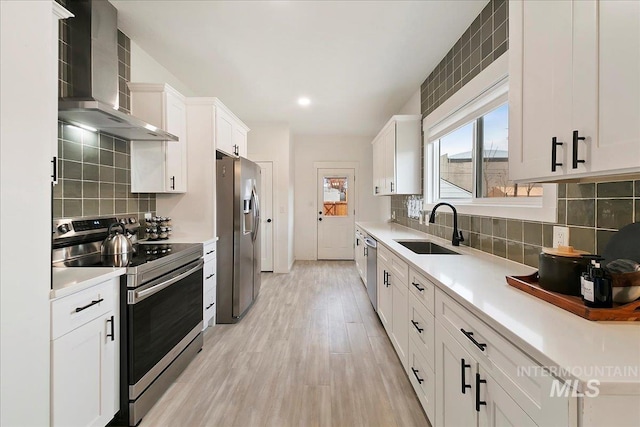 kitchen with light wood-type flooring, wall chimney exhaust hood, stainless steel appliances, sink, and white cabinetry