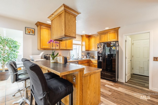 kitchen with a peninsula, light wood-type flooring, brown cabinets, black refrigerator with ice dispenser, and a kitchen bar