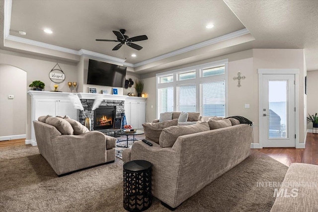 living room with a stone fireplace, ornamental molding, a raised ceiling, ceiling fan, and hardwood / wood-style floors