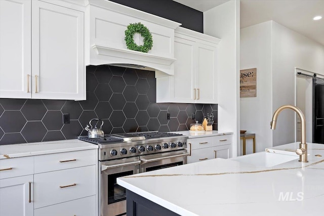 kitchen with a barn door, a sink, white cabinetry, backsplash, and range with two ovens