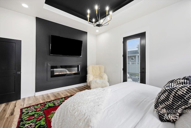 bedroom with a glass covered fireplace, a notable chandelier, recessed lighting, a tray ceiling, and wood finished floors