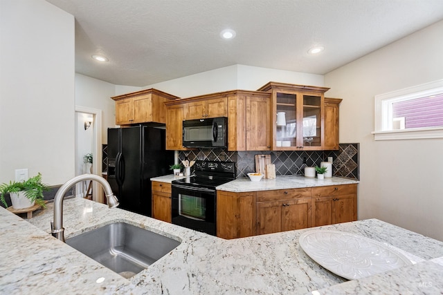 kitchen featuring brown cabinets, glass insert cabinets, a sink, light stone countertops, and black appliances