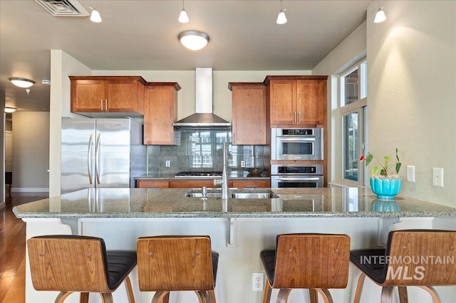 kitchen featuring backsplash, light stone counters, stainless steel appliances, wall chimney range hood, and a breakfast bar area